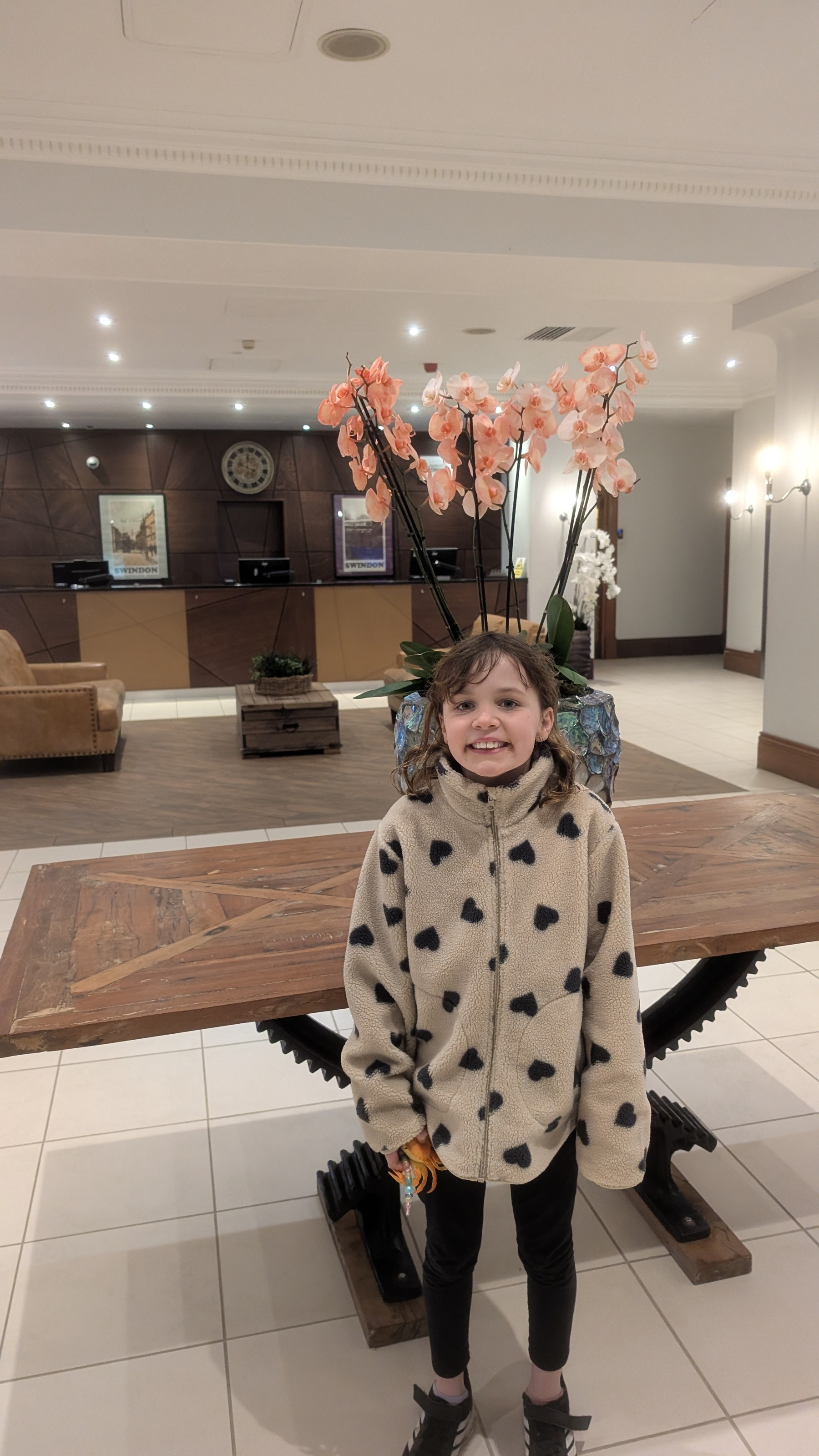 A child smiling in a hotel lobby stands in front of a table with potted orchids, with a reception desk in the background.