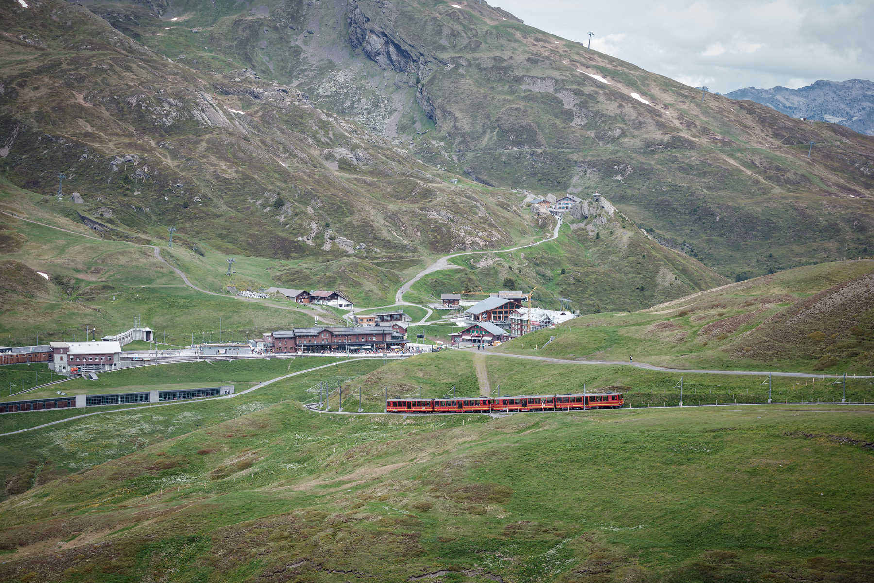 Un train rouge traverse un paysage montagneux verdoyant avec quelques bâtiments dispersés au pied des collines.