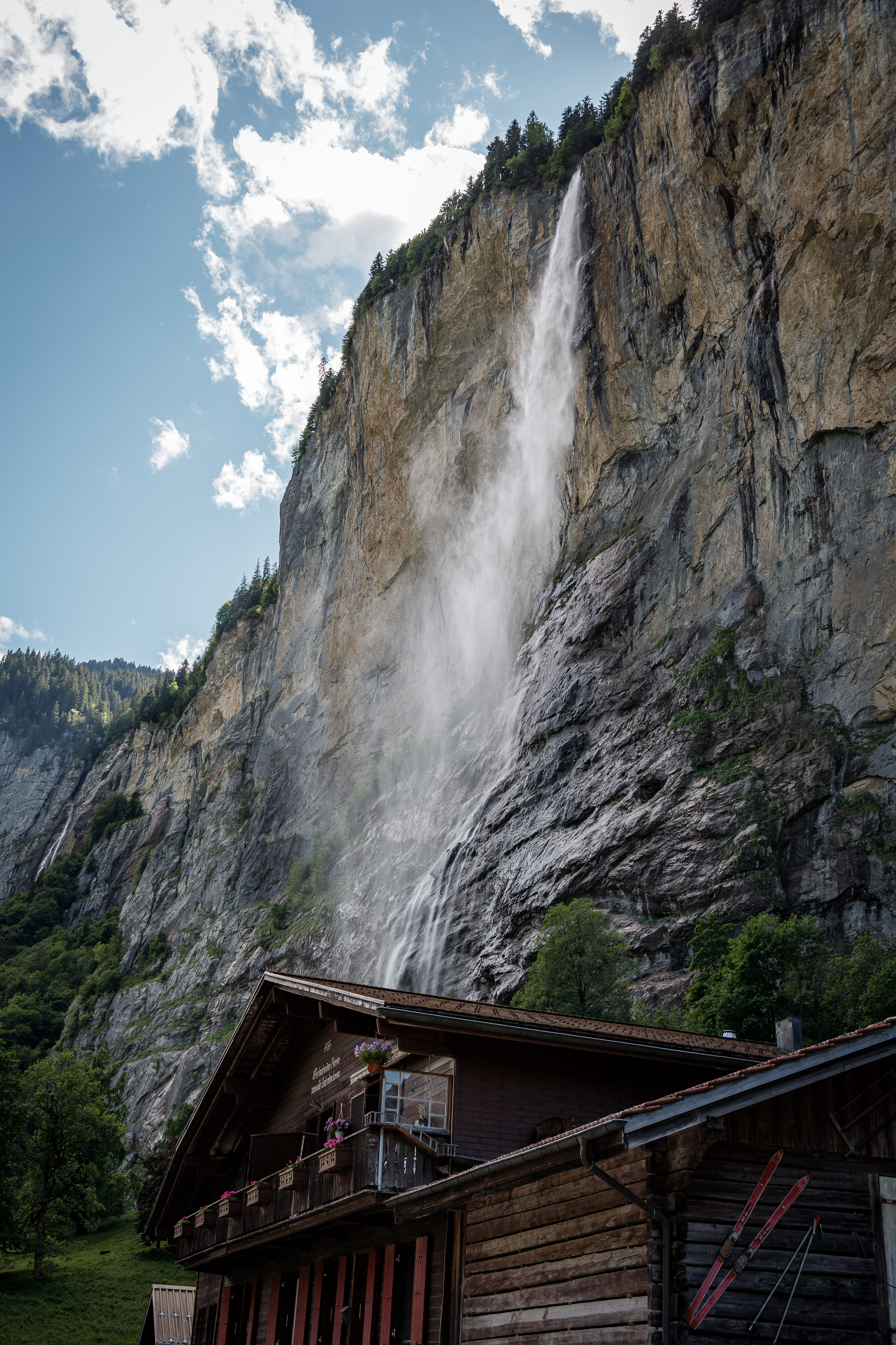 Une cascade spectaculaire dévale une falaise rocheuse derrière une maison en bois dans un paysage montagneux.