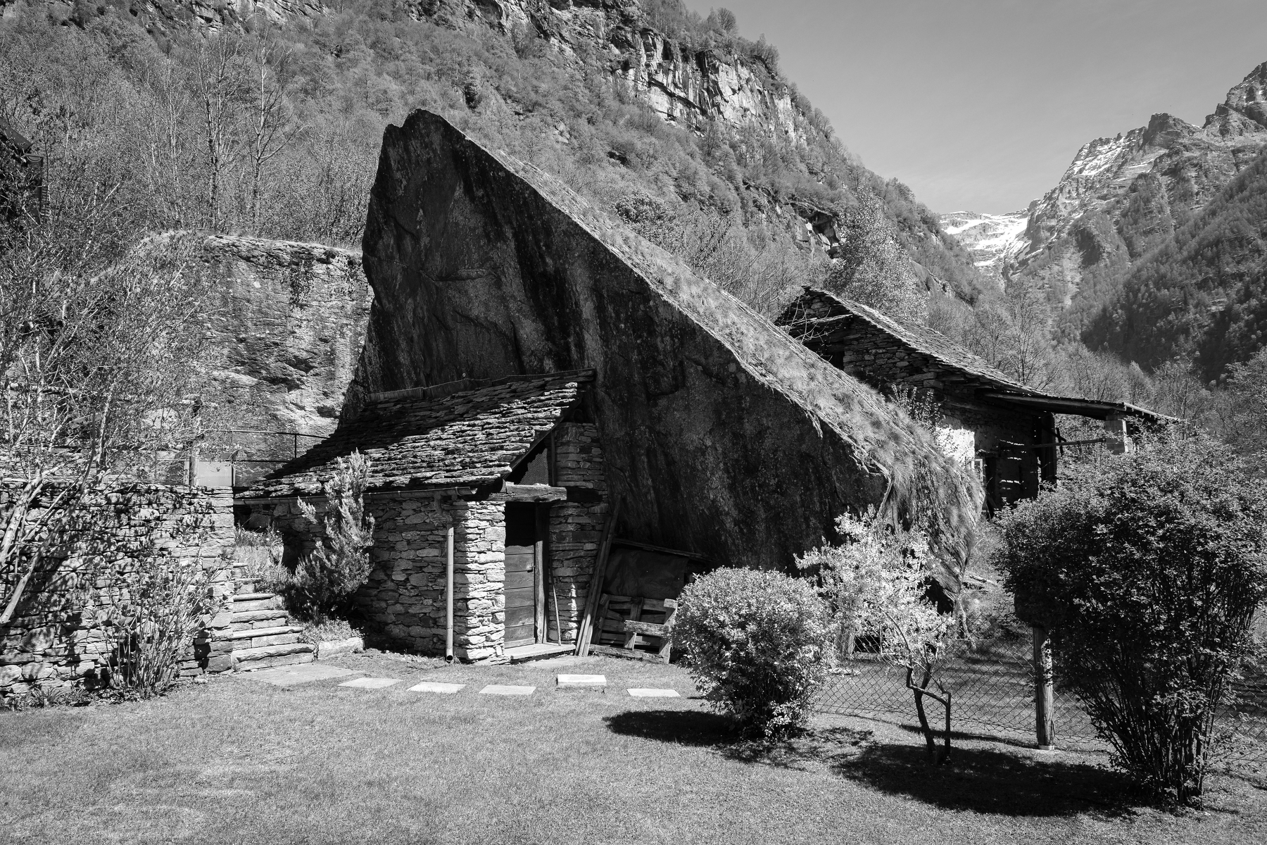 Auto-generated description: Une maison en pierre traditionnelle est nichée sous un grand roc incliné dans un paysage montagneux.