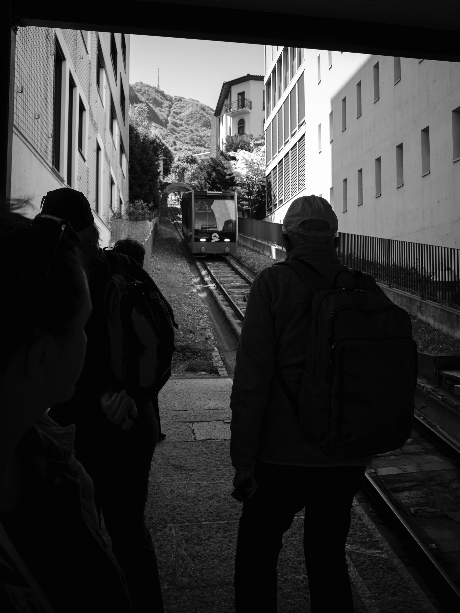 Auto-generated description: Des personnes attendent un funiculaire entrant dans une station située entre des bâtiments, avec des collines en arrière-plan.