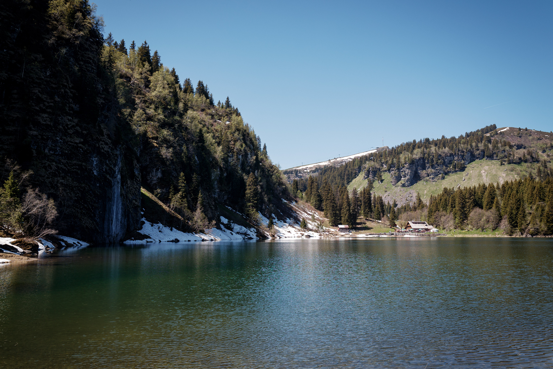 Auto-generated description: Un lac entouré de montagnes boisées avec quelques traces de neige et un chalet au loin.