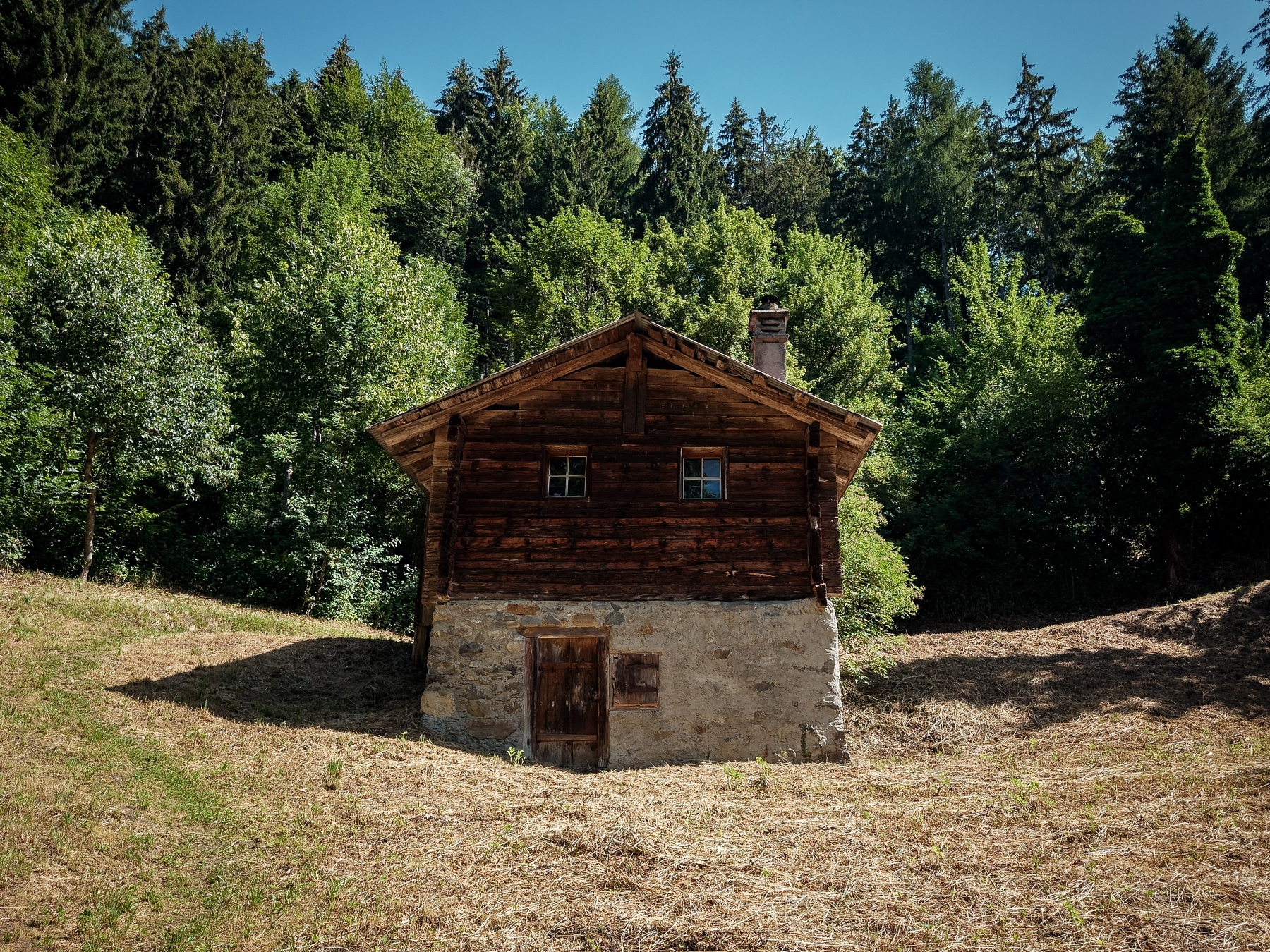 Auto-generated description: Une petite maison en bois et pierre se situe au milieu d'une clairière entourée d'arbres verts sous un ciel bleu.