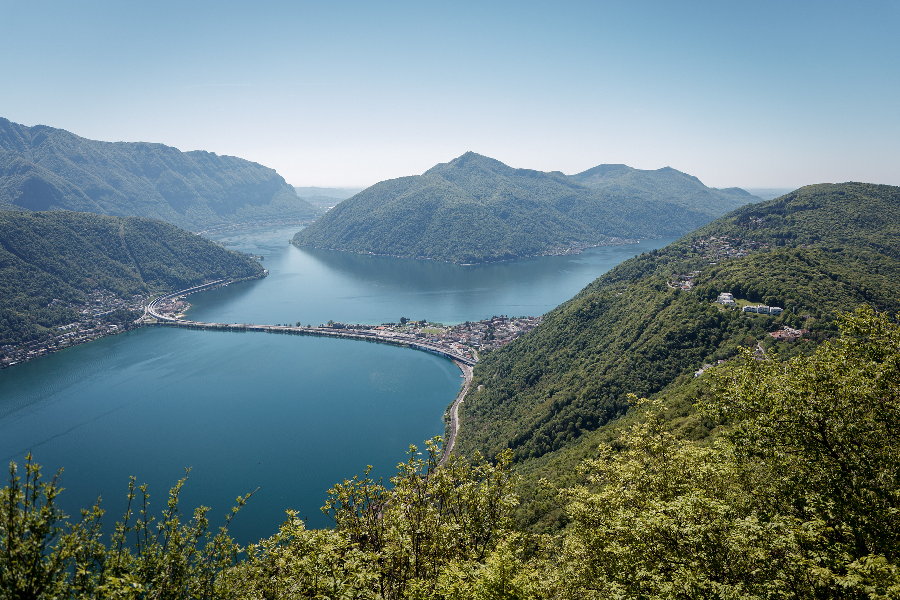 Auto-generated description: Un lac entouré de montagnes verdoyantes avec un pont traversant l'eau se dévoile sous un ciel bleu clair.