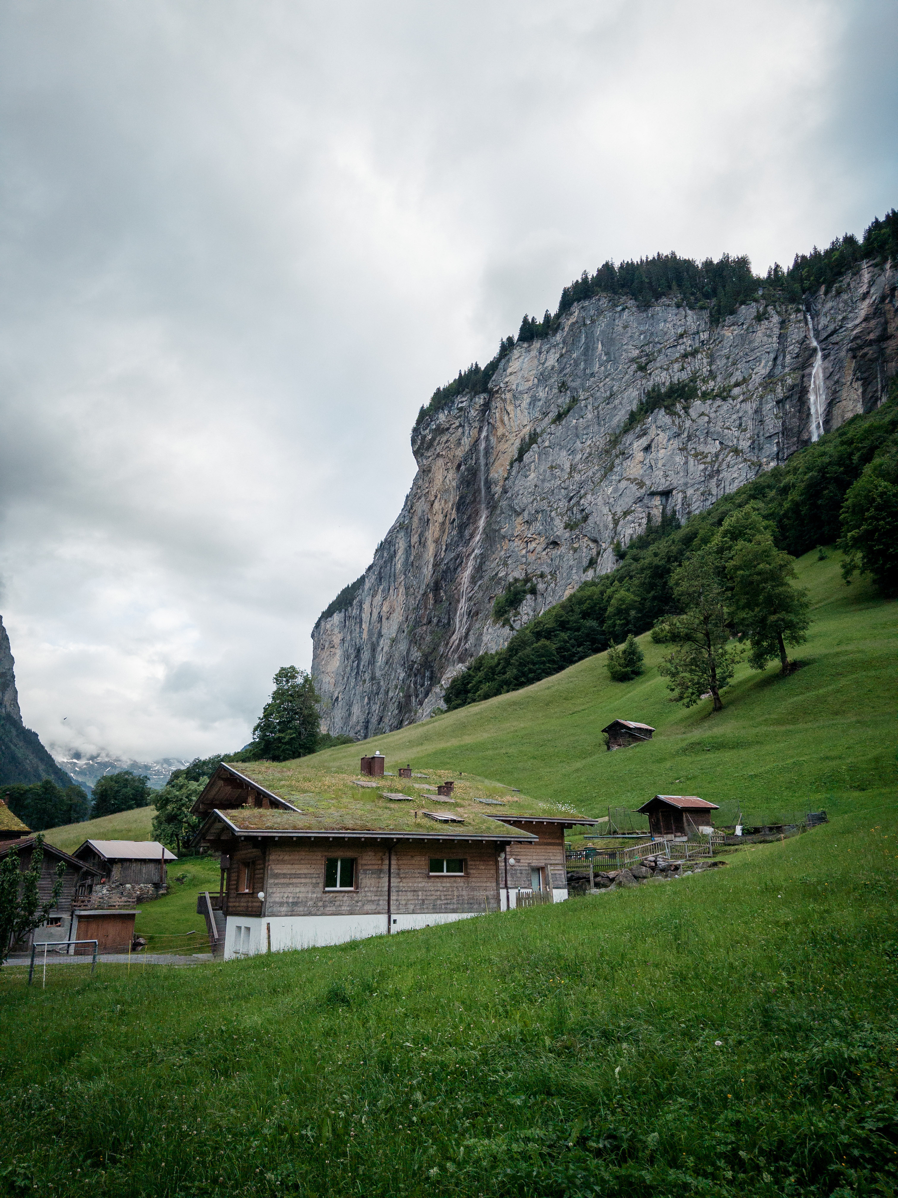 Une maison en bois avec un toit végétalisé est située au pied d'une falaise imposante entourée de prairies verdoyantes et quelques arbres sous un ciel nuageux.