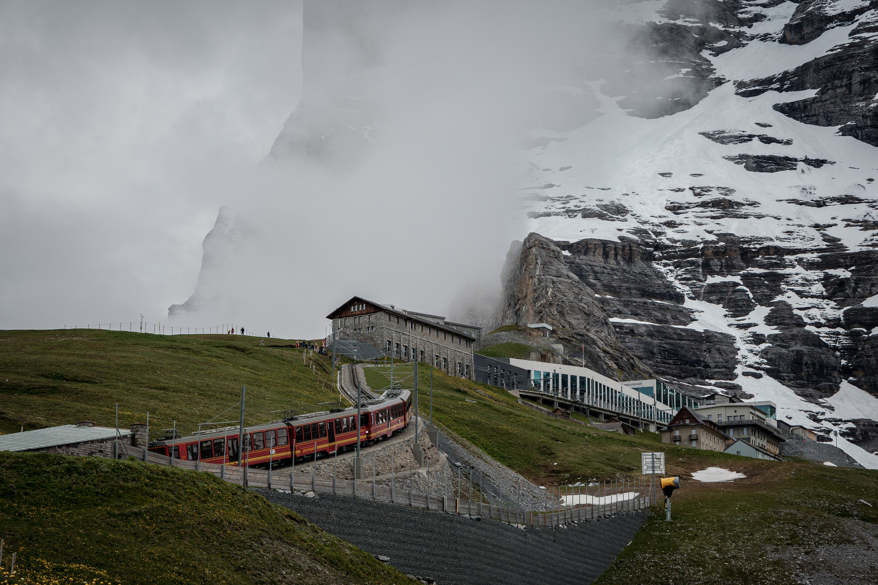 Un train rouge et blanc grimpe une montagne verdoyante avec des bâtiments en pierre et un sommet enneigé partiellement couvert de nuages en arrière-plan.