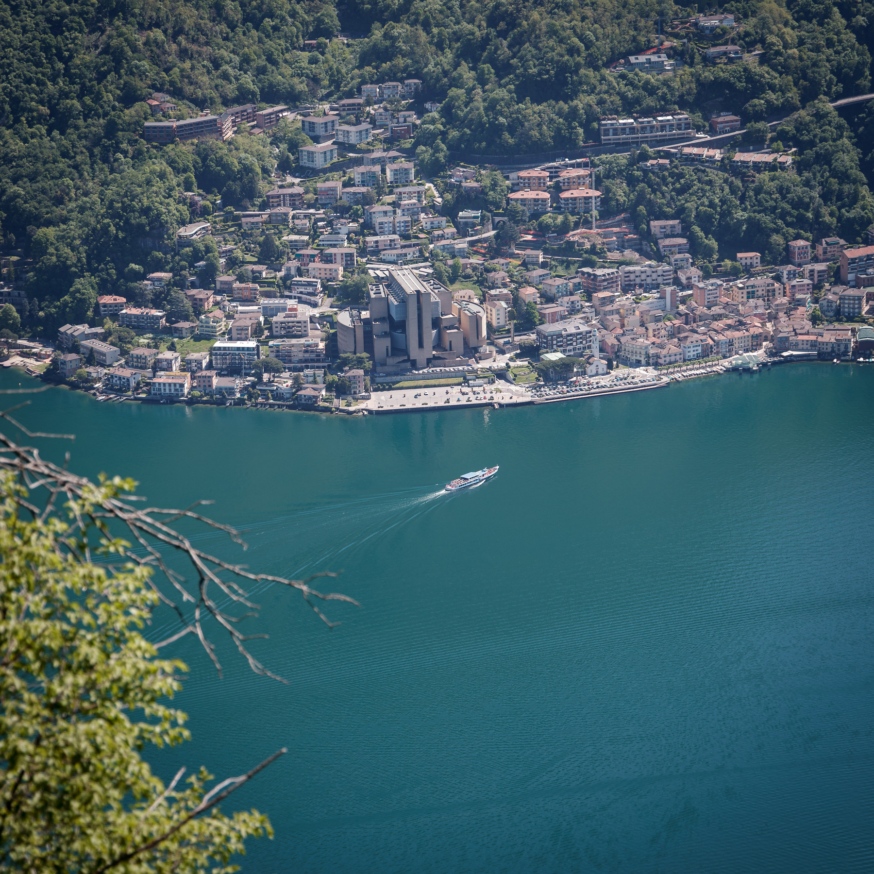 Auto-generated description: Une ville pittoresque bordant un lac avec un bateau naviguant au milieu de ses eaux, entourée de collines verdoyantes.