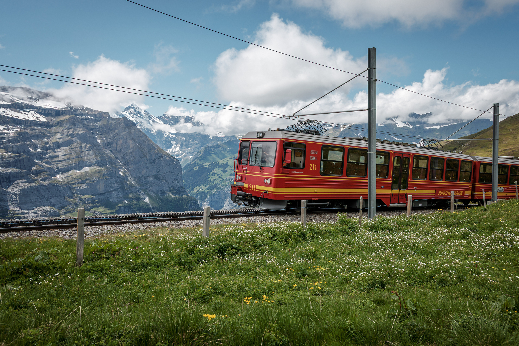 Un train rouge traverse un paysage montagneux avec des sommets enneigés et une prairie verdoyante.