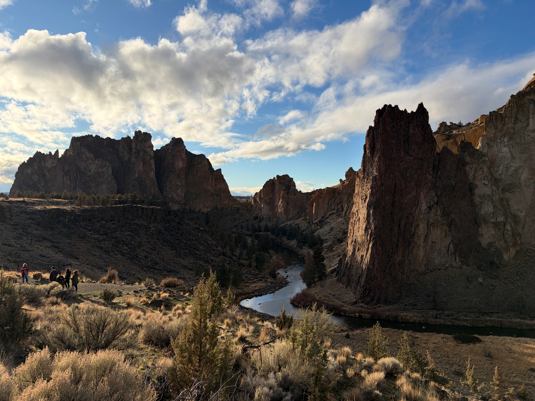 A winding river flows through a rocky canyon landscape under a partly cloudy sky with people on horseback in the foreground.