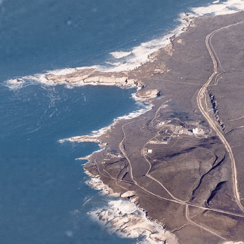 Auto-generated description: A coastal landscape features a rugged shoreline, small buildings, and winding pathways leading to the sea.