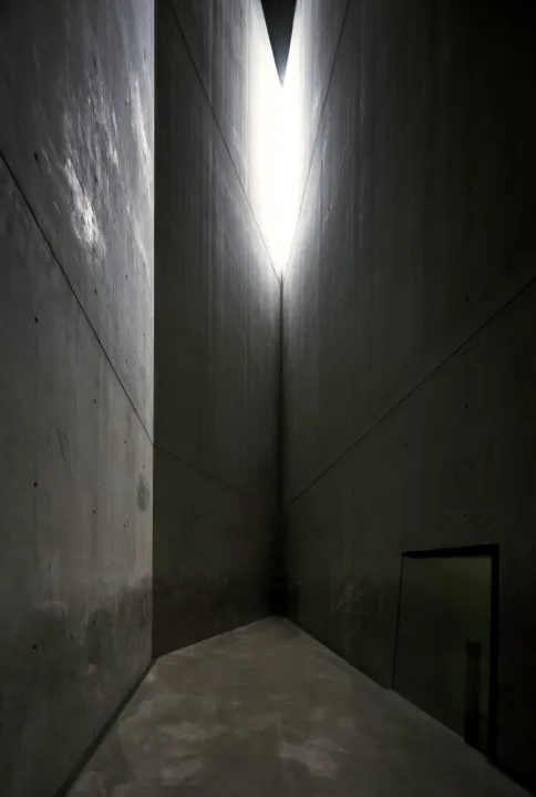 Auto-generated description: A narrow, triangular architectural space with concrete walls and a beam of light shining from above.