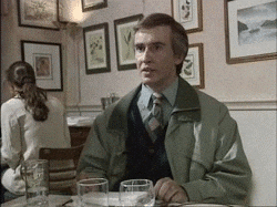 Auto-generated description: Alan Partridge sits at a table in a restaurant and shrugs.