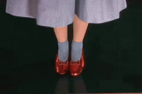 Auto-generated description: Judy Garland wearing a blue gingham dress stands with feet together, tapping together her red ruby slippers.