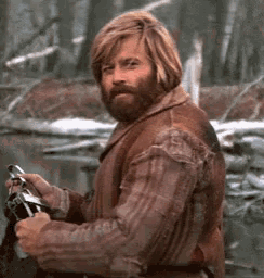 Auto-generated description: A bearded man in rustic attire, holding an object, looks over his shoulder in a woodland setting.
