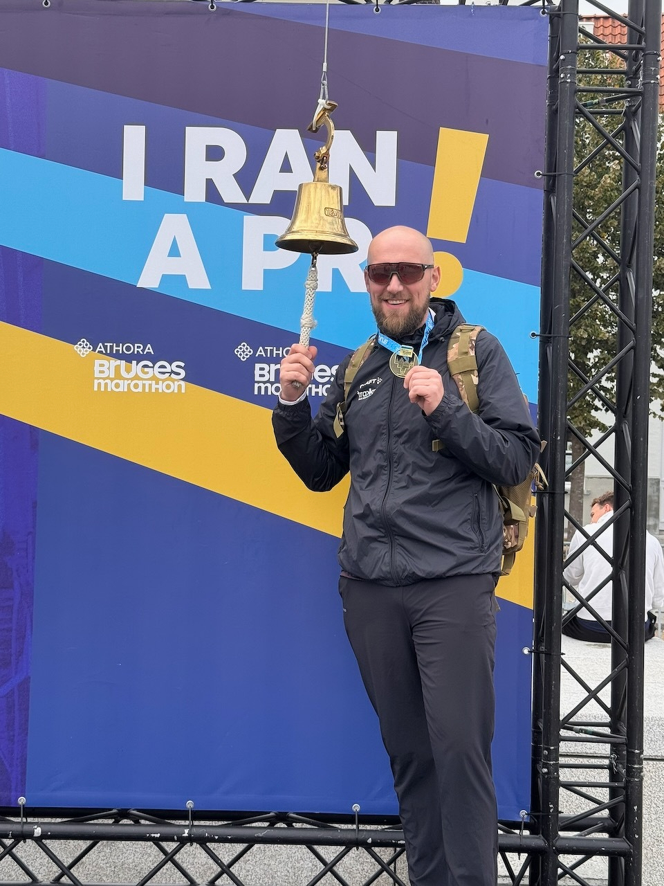 Auto-generated description: Un homme souriant portant une médaille et des lunettes de soleil sonne une cloche devant une bannière indiquant I RAN A PR! lors du marathon d'Athora Bruges.