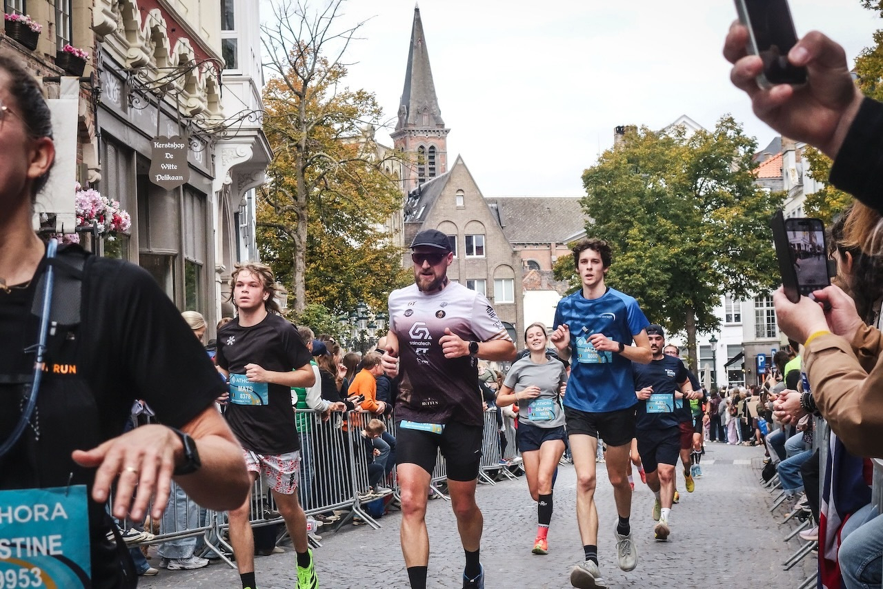 Auto-generated description: Des coureurs participent à une course urbaine tout en étant encouragés par des spectateurs le long d'une rue pavée bordée de bâtiments historiques.