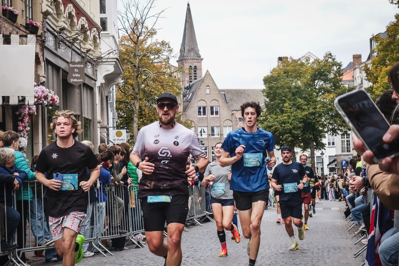 Auto-generated description: Des coureurs participent à une course en ville devant un public enthousiaste, avec des bâtiments historiques en arrière-plan.