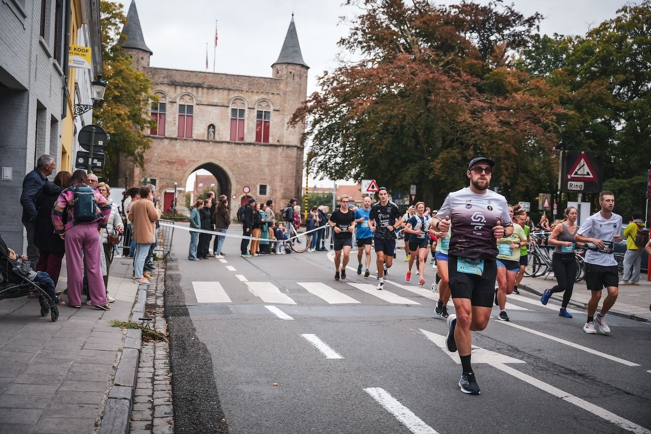Auto-generated description: Un groupe de coureurs participe à une course dans une ville avec des spectateurs le long du trottoir et un bâtiment historique en arrière-plan.