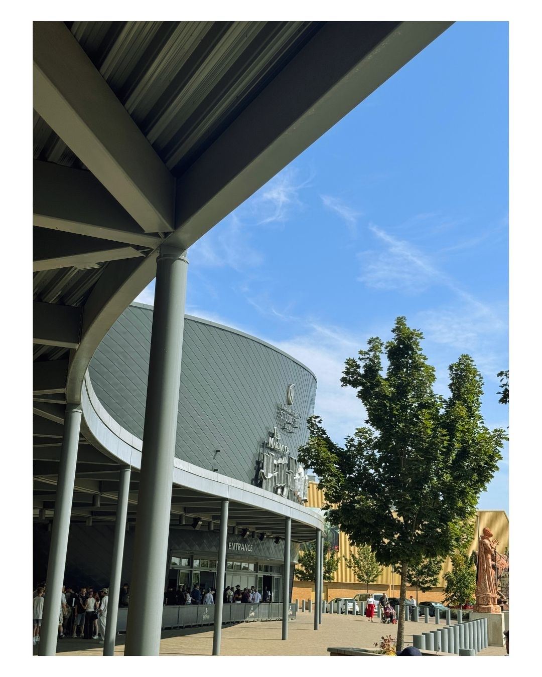 Auto-generated description: Une file de personnes attend sous une grande structure moderne en métal devant un bâtiment arrondi avec un ciel bleu et quelques arbres à proximité.