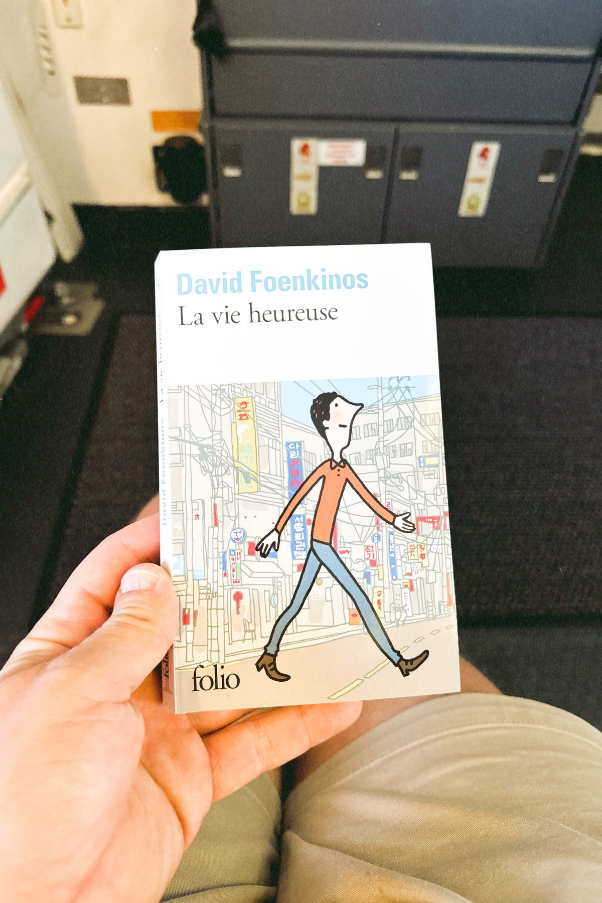 Auto-generated description: Une personne tient le livre La vie heureuse de David Foenkinos, publié chez folio, avec un fond flou semblant être l'intérieur d'un moyen de transport.