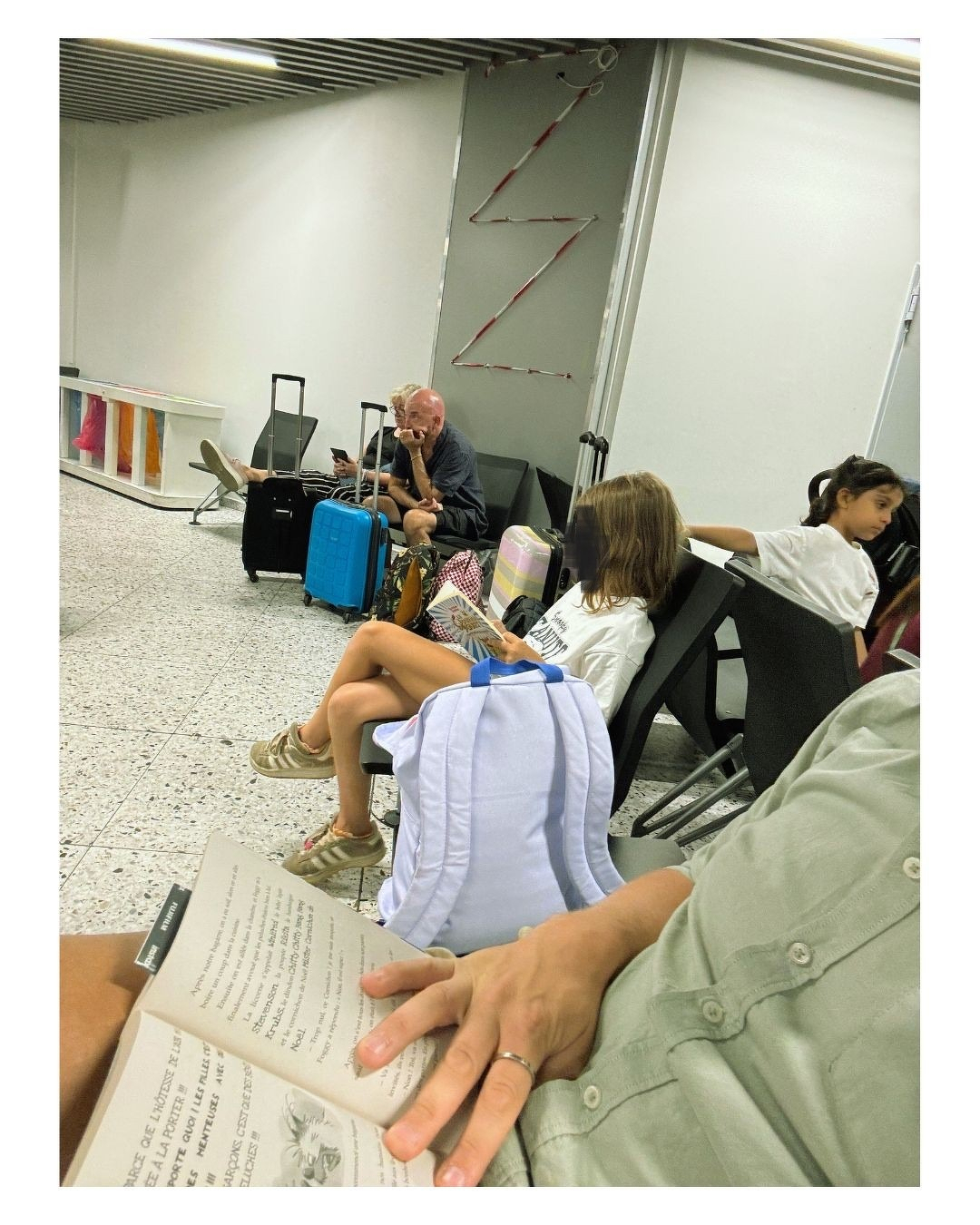 Auto-generated description: Plusieurs personnes, dont des enfants et des adultes, attendent assises dans une salle d'attente avec des valises, tandis qu'une personne au premier plan lit un livre.