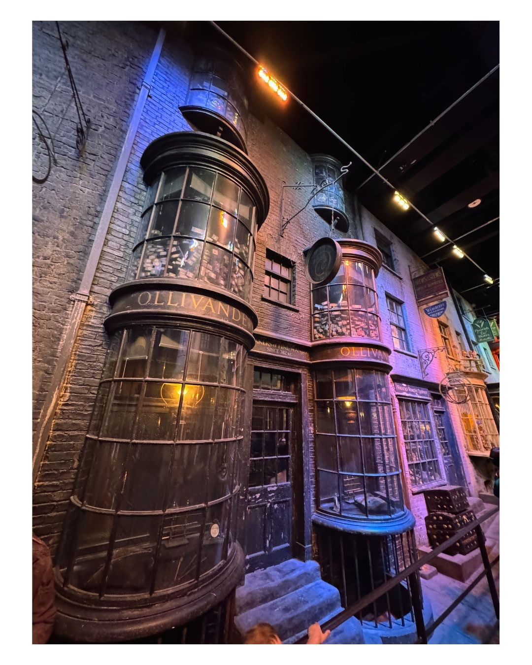Auto-generated description: Une façade ancienne avec des vitrines arrondies affichant le nom OLLIVANDER, probablement une boutique de baguettes magiques inspirée de l'univers de Harry Potter.