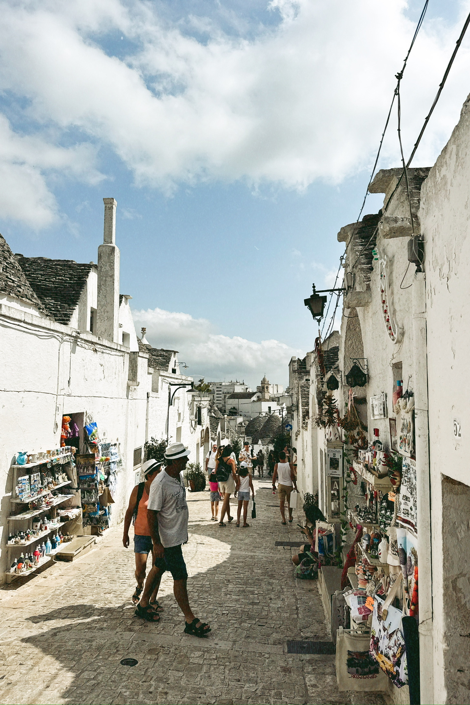 Auto-generated description: Une ruelle pavée animée bordée de bâtiments blancs avec des touristes qui explorent des boutiques d'artisanat sous un ciel partiellement nuageux.