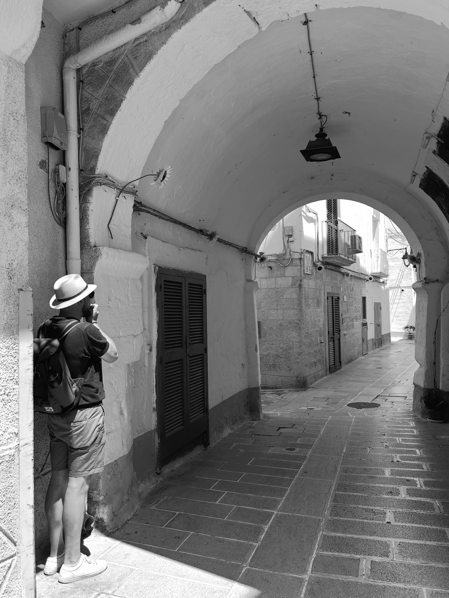 Auto-generated description: Un homme portant un chapeau et un sac à dos prend une photo dans une ruelle étroite sous un arc voûté.