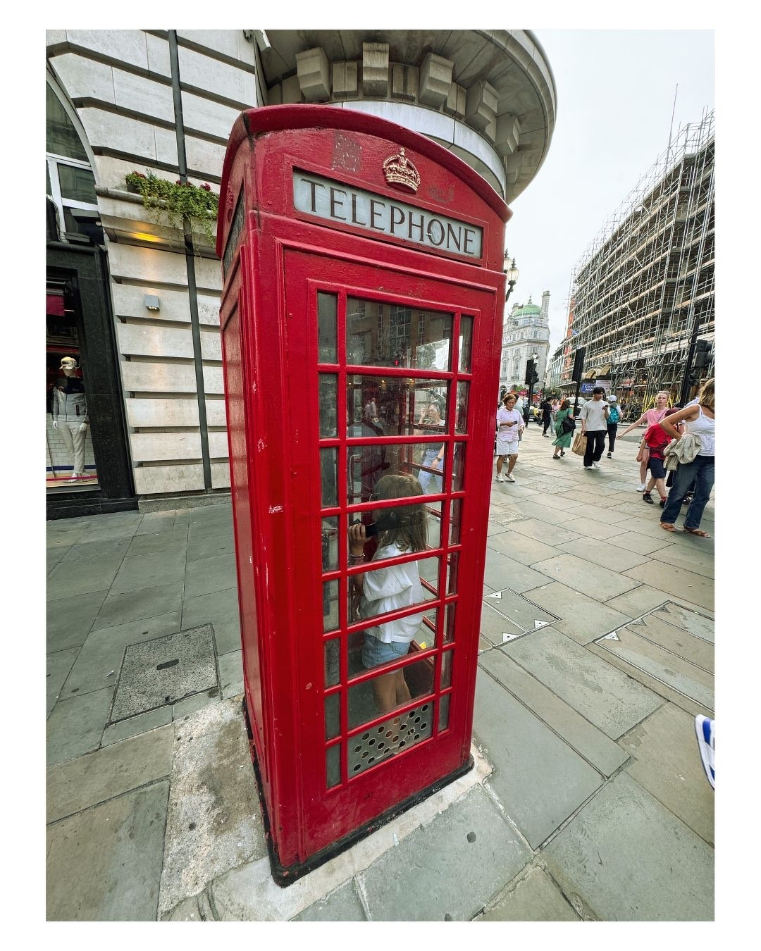 Auto-generated description: Une cabine téléphonique rouge traditionnelle britannique est située sur un trottoir animé avec une personne à l'intérieur.