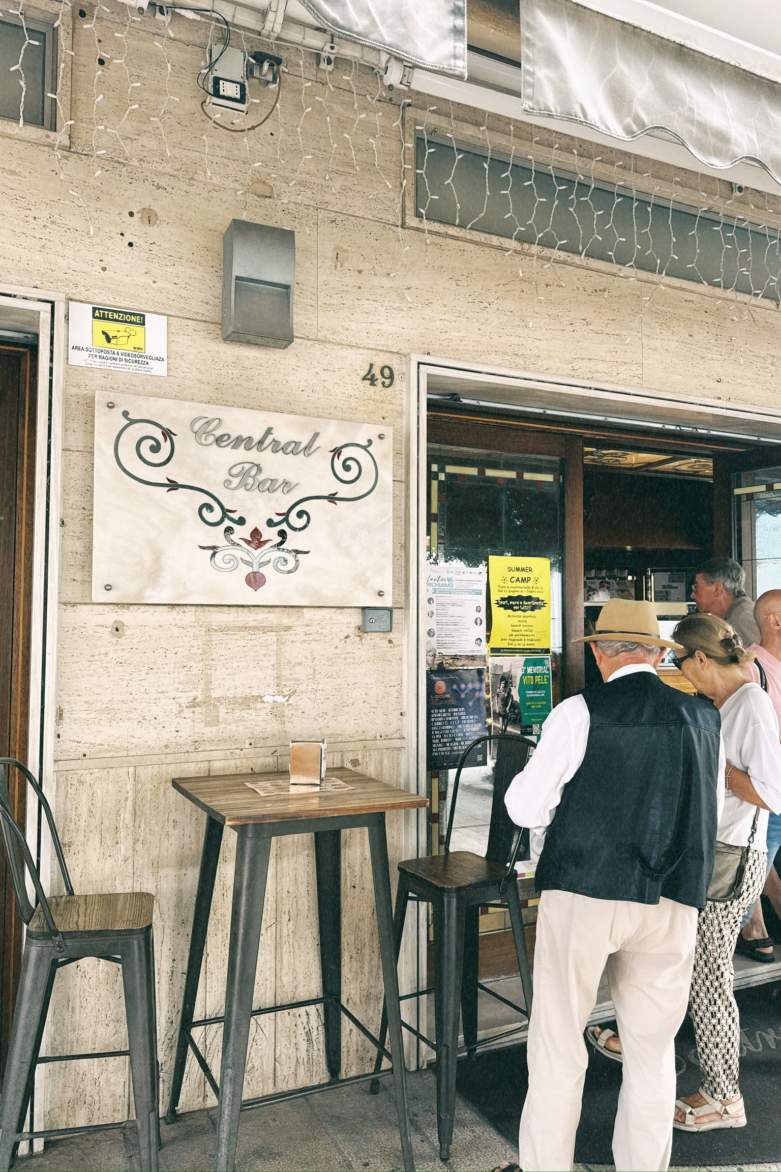 Auto-generated description: Des personnes attendent à l'entrée d'un bar nommé Central Bar orné de décorations murales et entouré de chaises hautes.