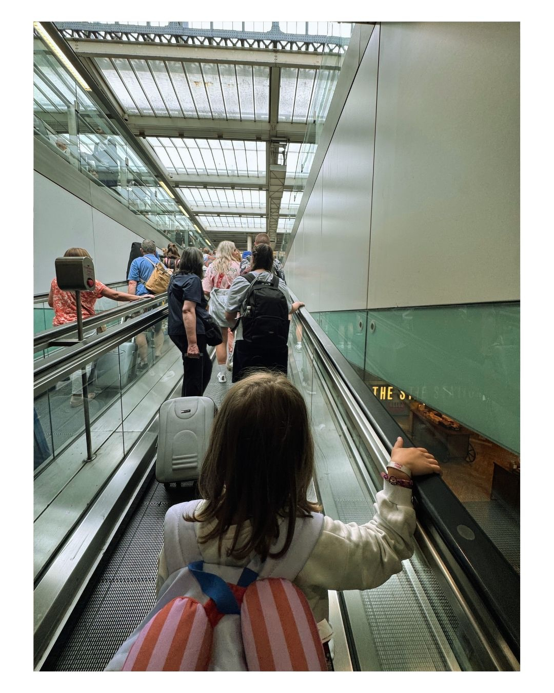 Auto-generated description: Une fille avec un sac à dos rayé monte un escalator entourée d'autres personnes dans un espace lumineux et moderne.