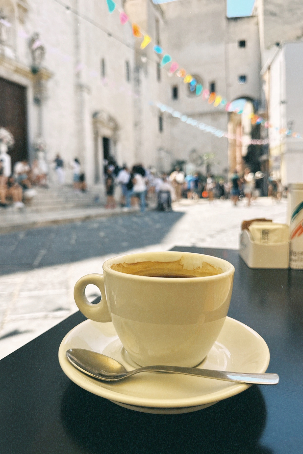 Auto-generated description: Une tasse de café sur une table en terrasse avec une rue pavée animée en arrière-plan.