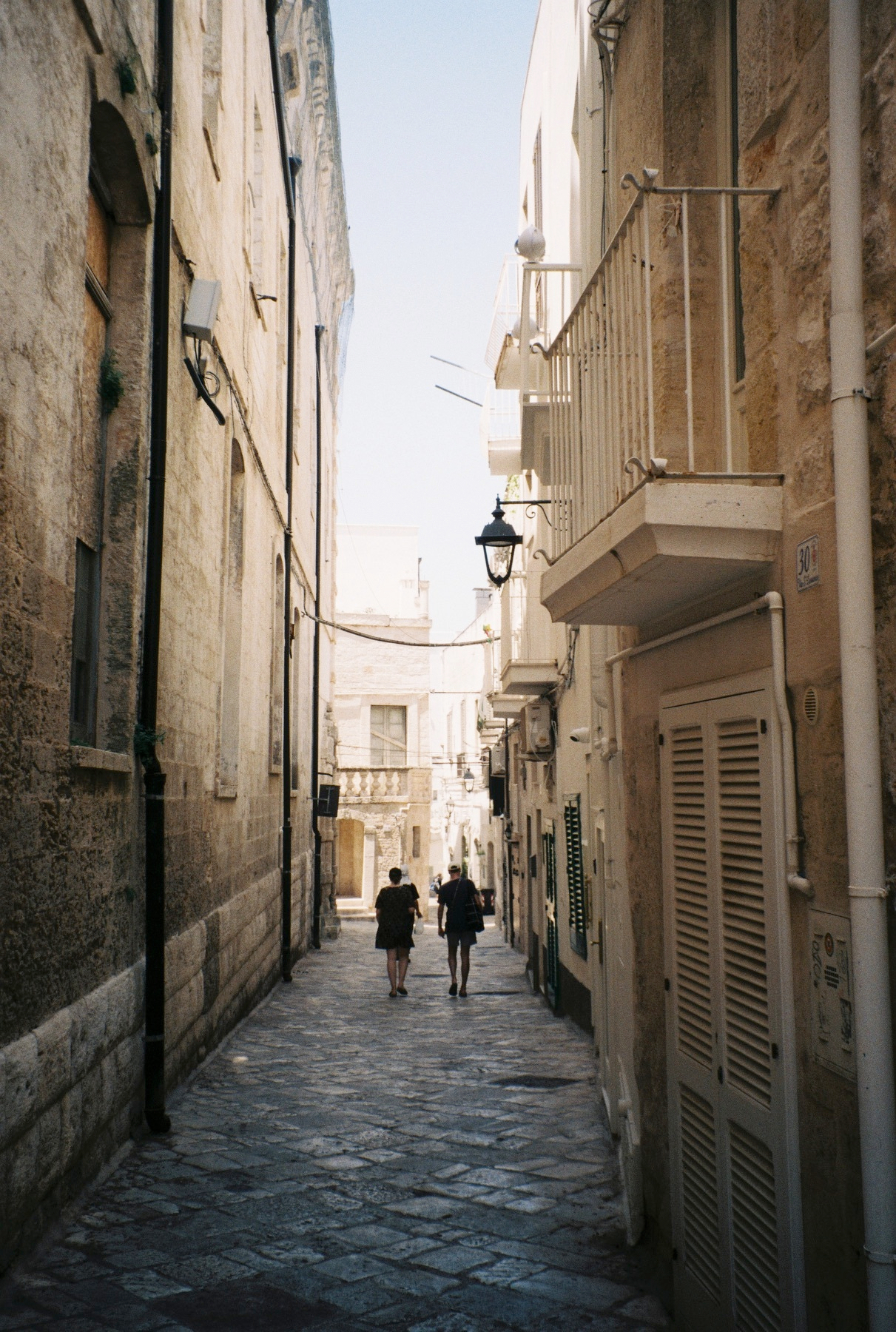 Auto-generated description: Deux personnes marchent dans une ruelle étroite bordée de bâtiments en pierre sous un ciel clair.