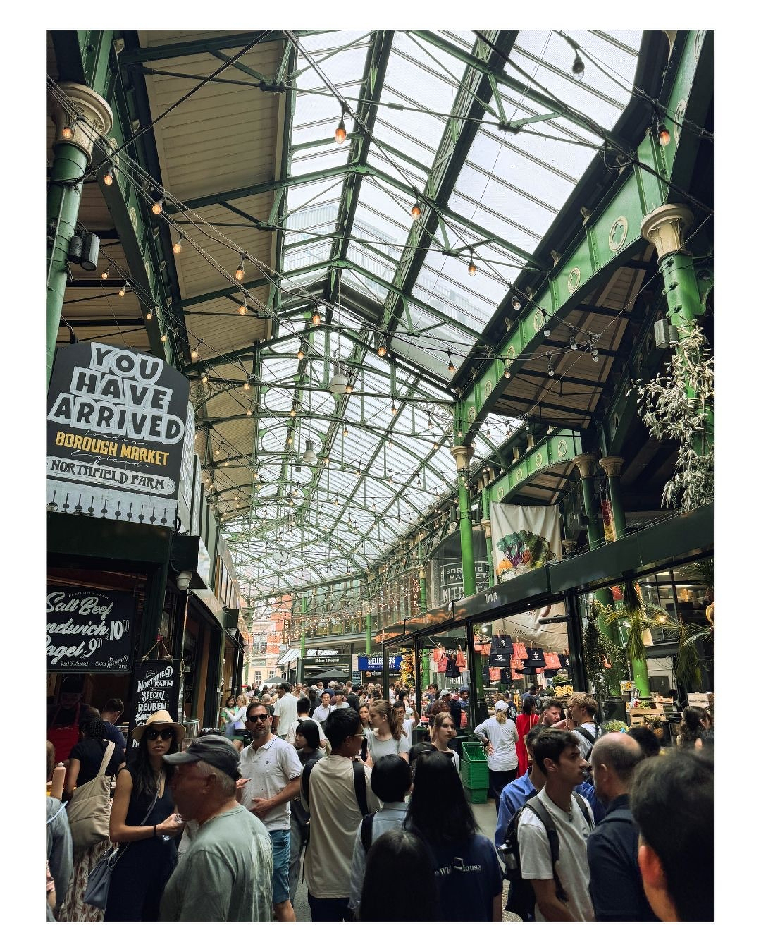 Auto-generated description: Une foule dense de personnes se promène sous une grande verrière en métal vert dans un marché couvert animé.