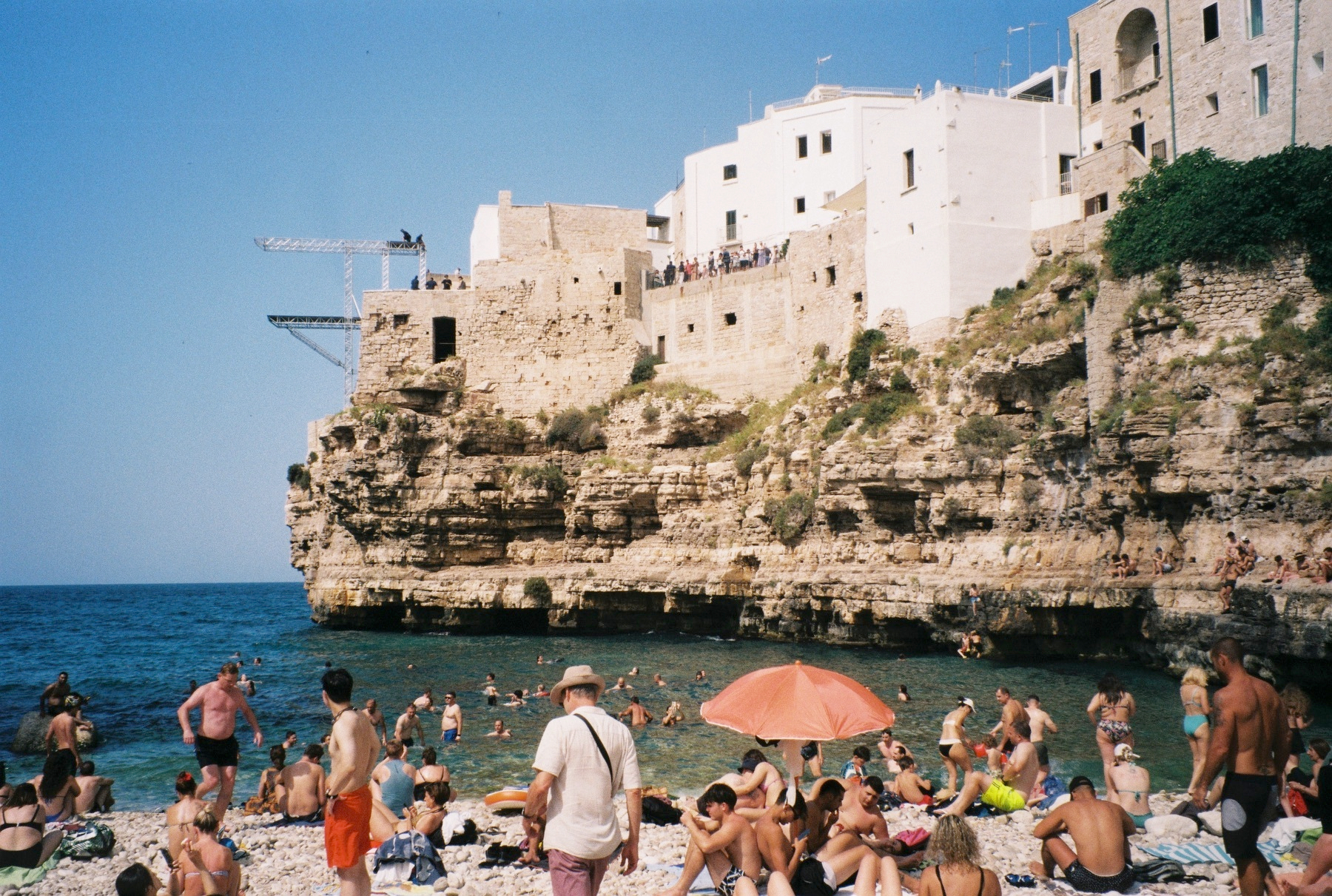 Auto-generated description: Une plage rocheuse animée est bordée par une falaise avec des maisons blanches surplombant la mer, où de nombreuses personnes se baignent et se détendent sous un ciel bleu clair.