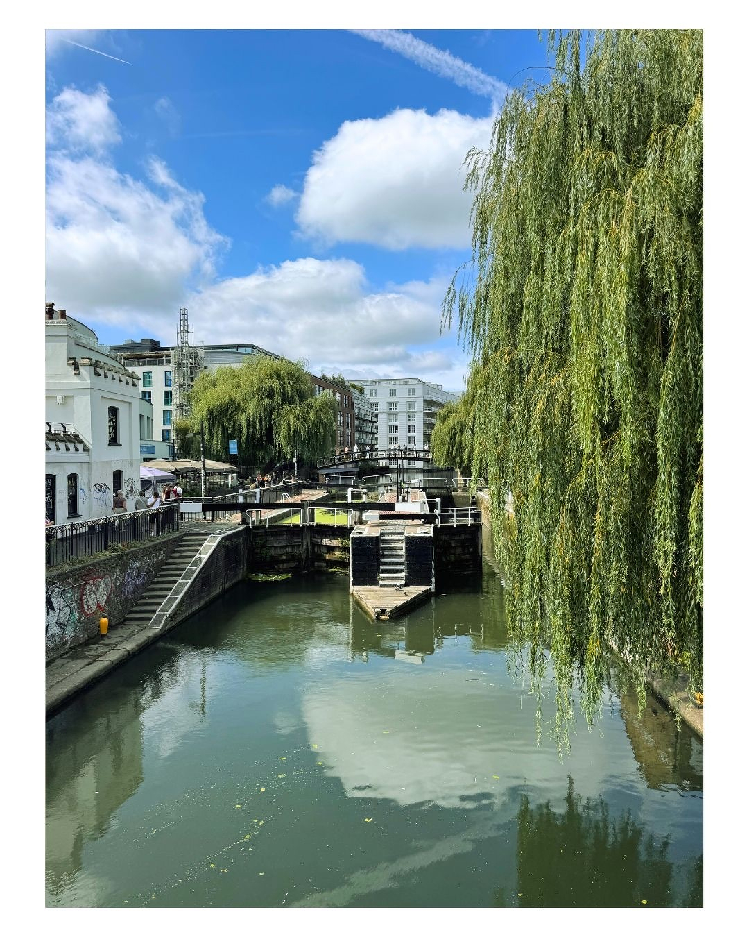 Auto-generated description: Un canal bordé d'arbres et de bâtiments, avec un ciel bleu partiellement nuageux reflété dans l'eau.