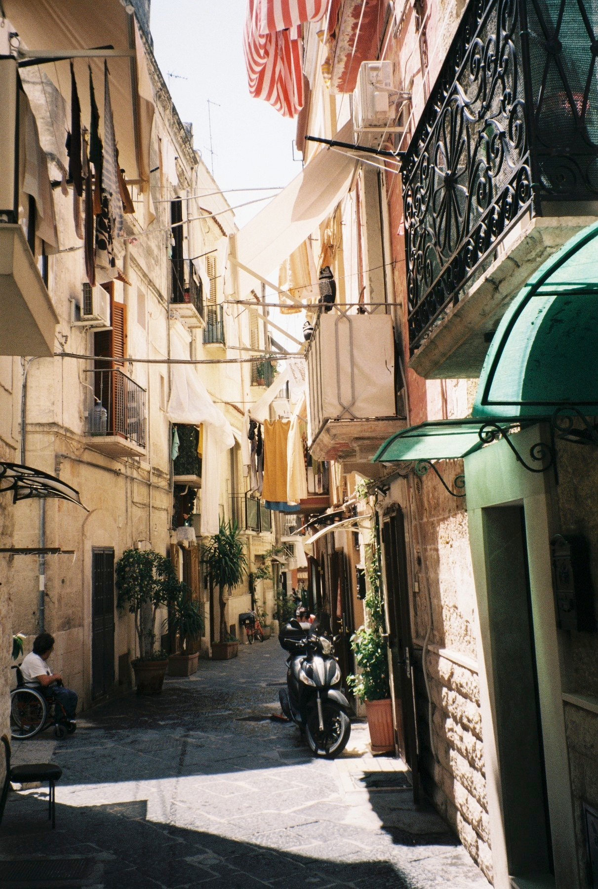 Auto-generated description: Une ruelle étroite et ensoleillée avec des vêtements suspendus, des plantes en pot, un scooter garé et une personne en fauteuil roulant.