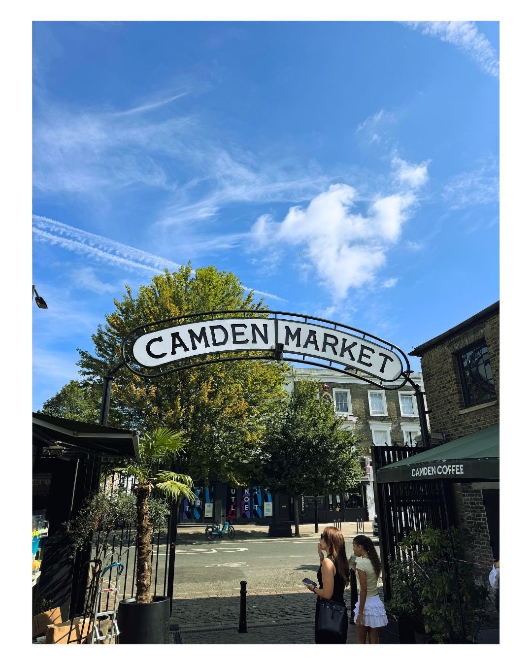 Auto-generated description: L'entrée du marché de Camden à Londres est visible sous un ciel bleu avec quelques nuages, avec deux personnes se tenant à proximité.