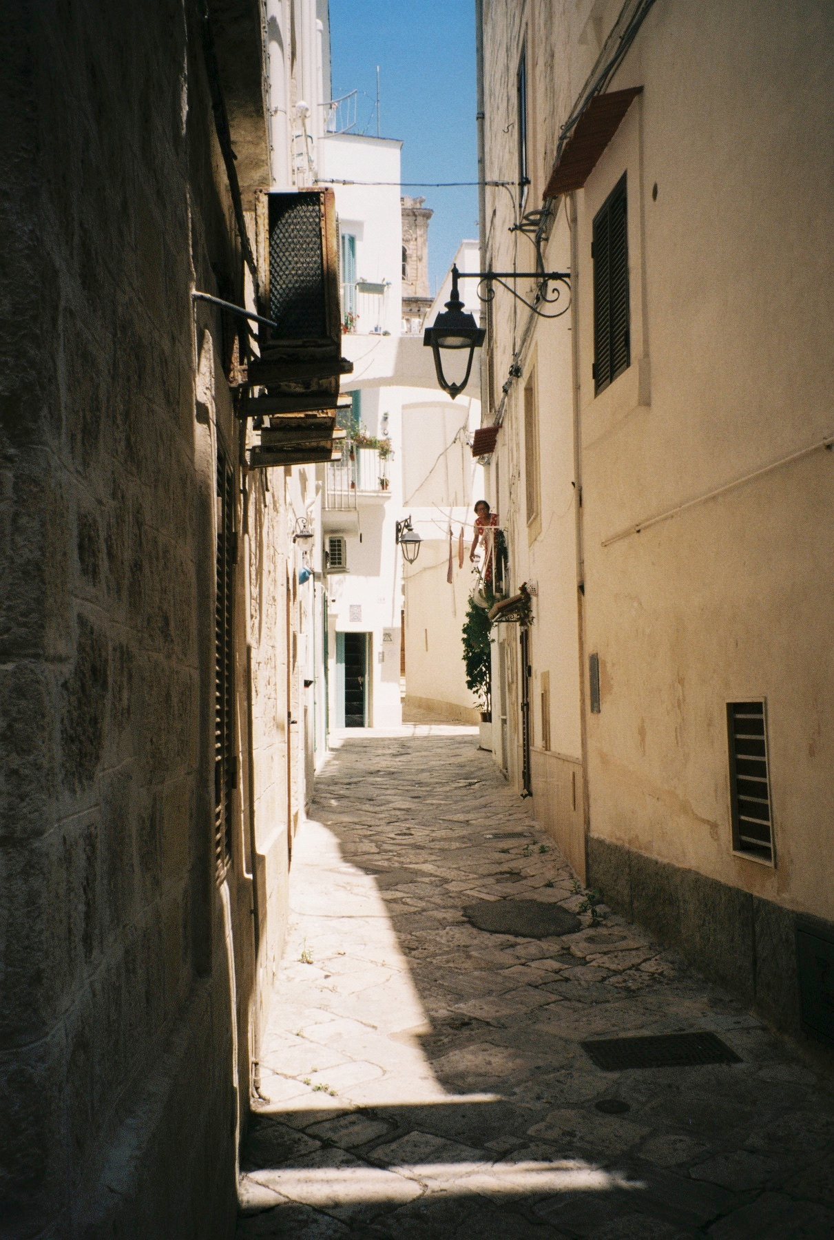 Auto-generated description: Une ruelle étroite pavée est bordée de bâtiments aux murs clairs avec quelques lampes suspendues et une personne debout sur un balcon.