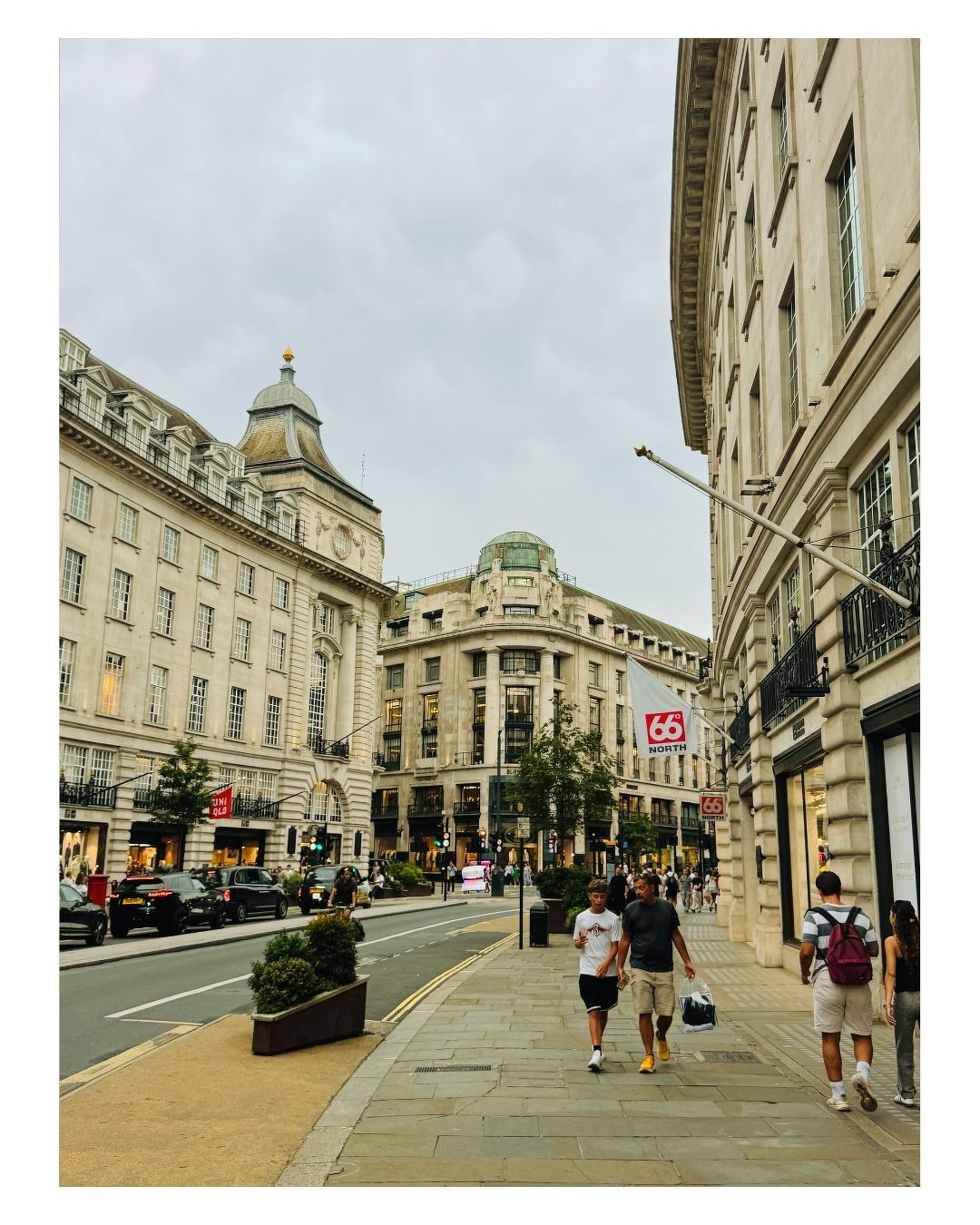 Auto-generated description: Une rue animée d'une ville européenne montre des bâtiments classiques, des piétons et des voitures sous un ciel nuageux.