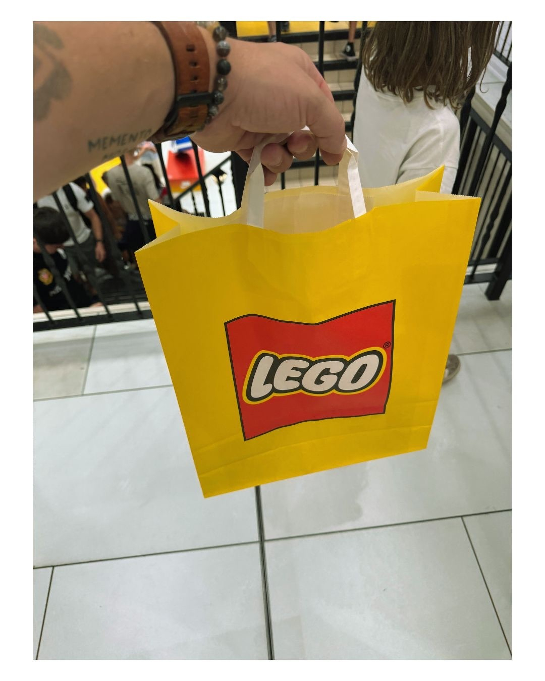 Auto-generated description: Une main tient un sac jaune portant le logo LEGO dans un environnement intérieur avec des personnes en arrière-plan.