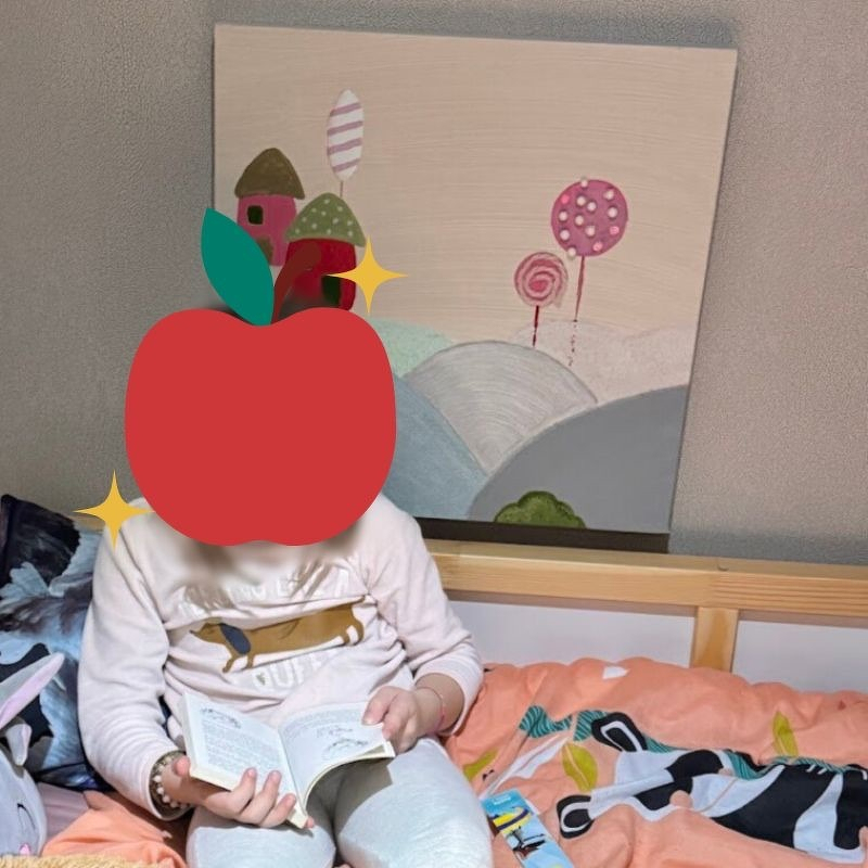 Auto-generated description: Un enfant est assis sur un lit en train de lire un livre avec un tableau coloré accroché au mur derrière lui.
