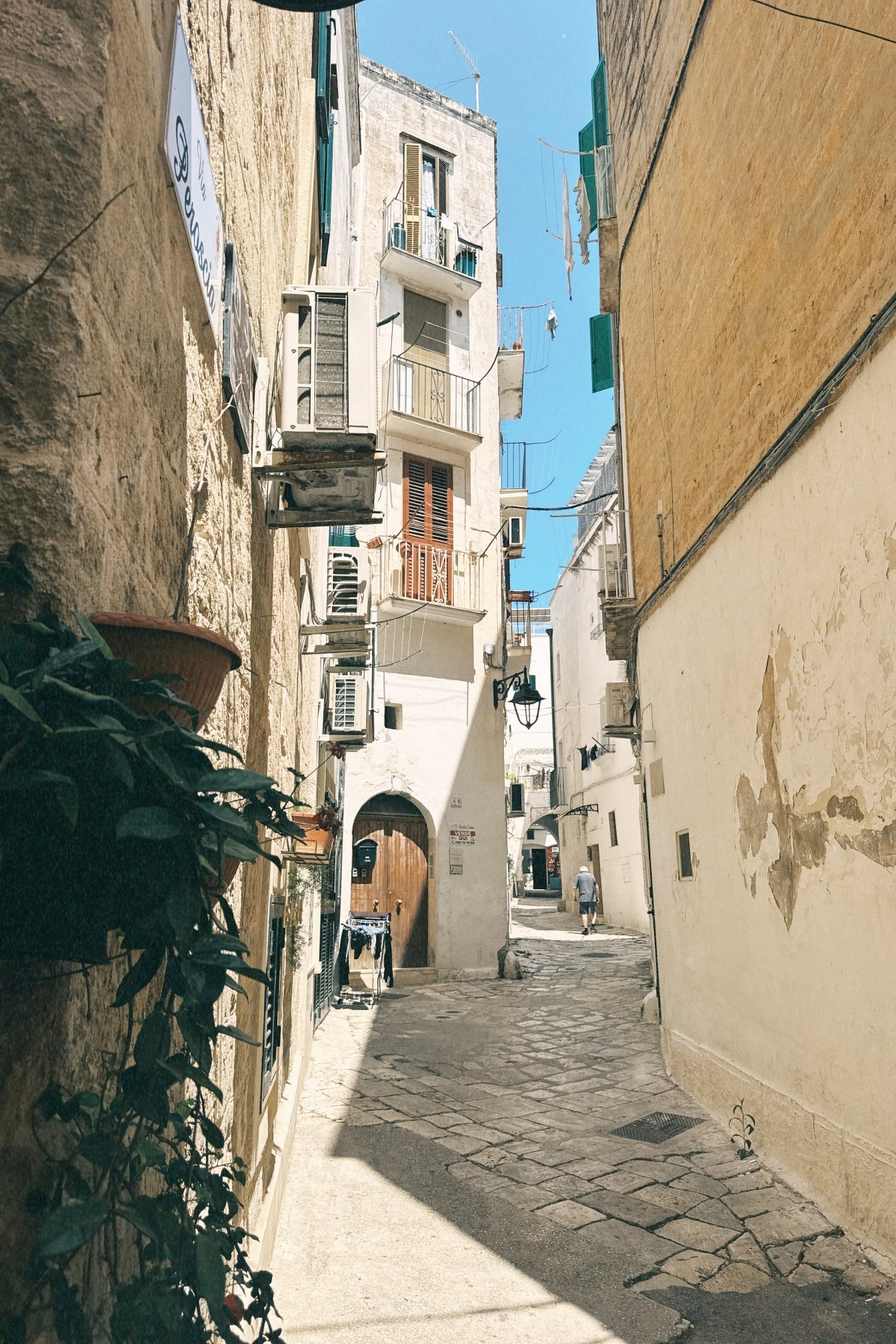 Auto-generated description: Une ruelle étroite bordée de bâtiments anciens avec des fenêtres, des balcons, des climatiseurs et des vêtements suspendus, sous un ciel bleu clair.