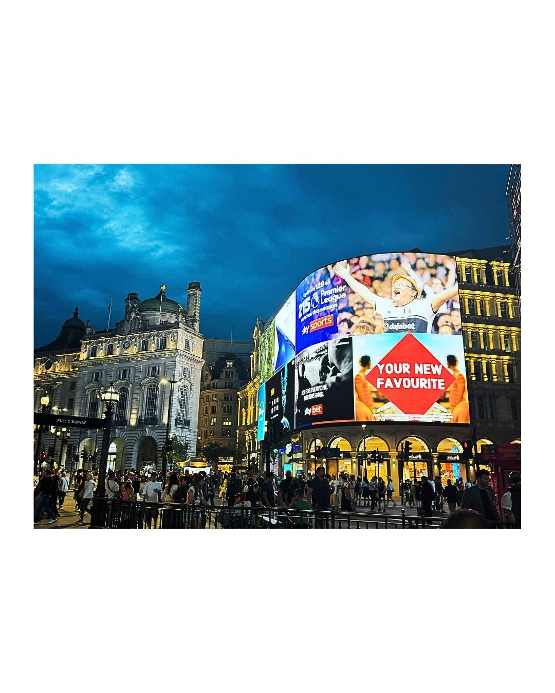 Auto-generated description: Une foule se rassemble dans une place urbaine animée en soirée, illuminée par de grands écrans publicitaires colorés.