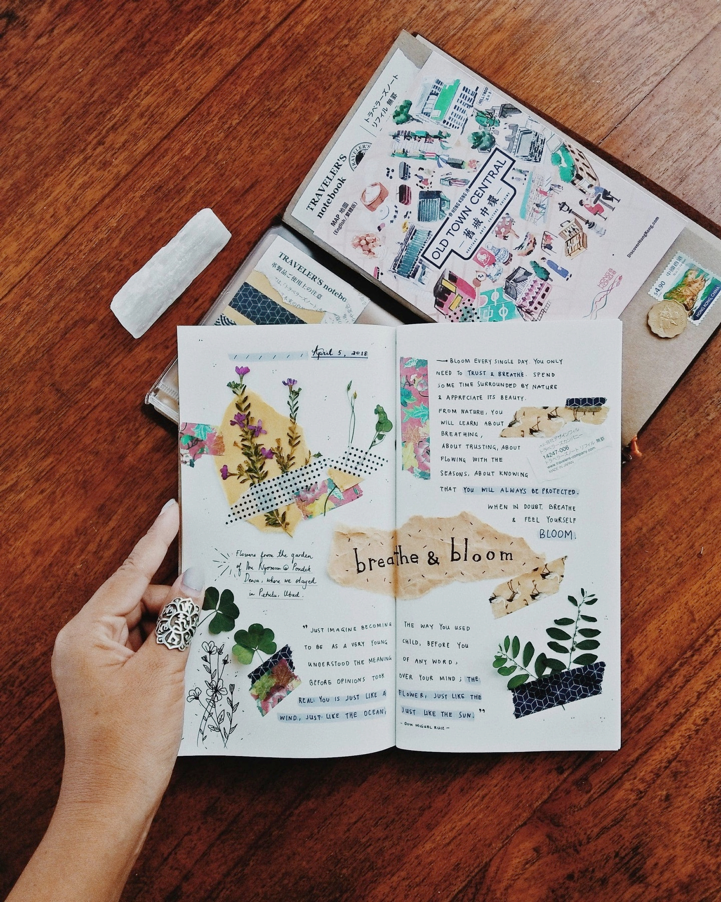 Auto-generated description: Un carnet de notes décoré avec des collages floraux, des citations manuscrites et des décorations en masking tape repose sur une table en bois, accompagné de deux livres et d'un objet blanc.
