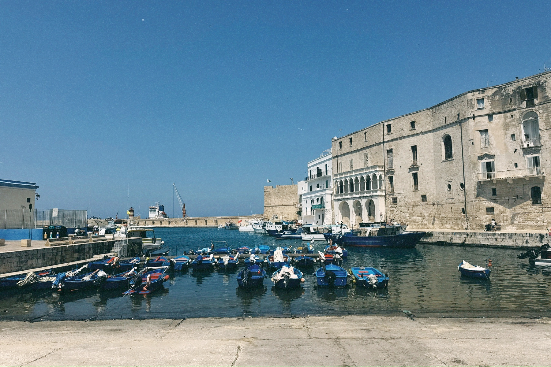 Auto-generated description: Un port calme avec plusieurs petites barques amarrées près d'un quai, bordé par de vieux bâtiments en pierre sous un ciel bleu clair.