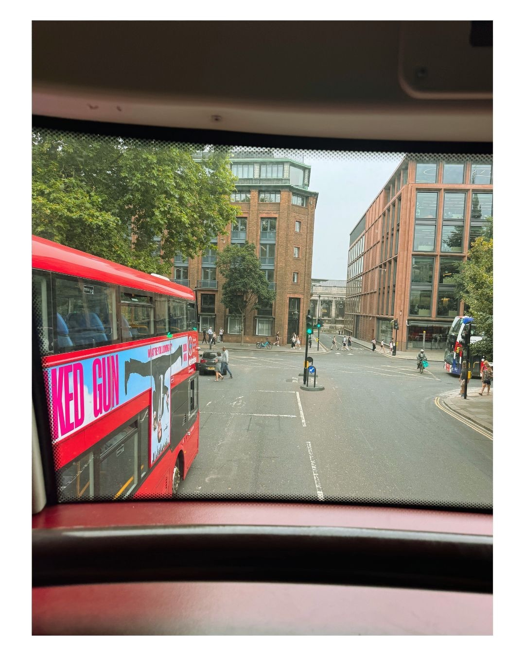 Auto-generated description: Un bus rouge avec une publicité est vu à travers la fenêtre d'un autre véhicule dans une rue urbaine animée, entourée de bâtiments modernes et d'arbres.