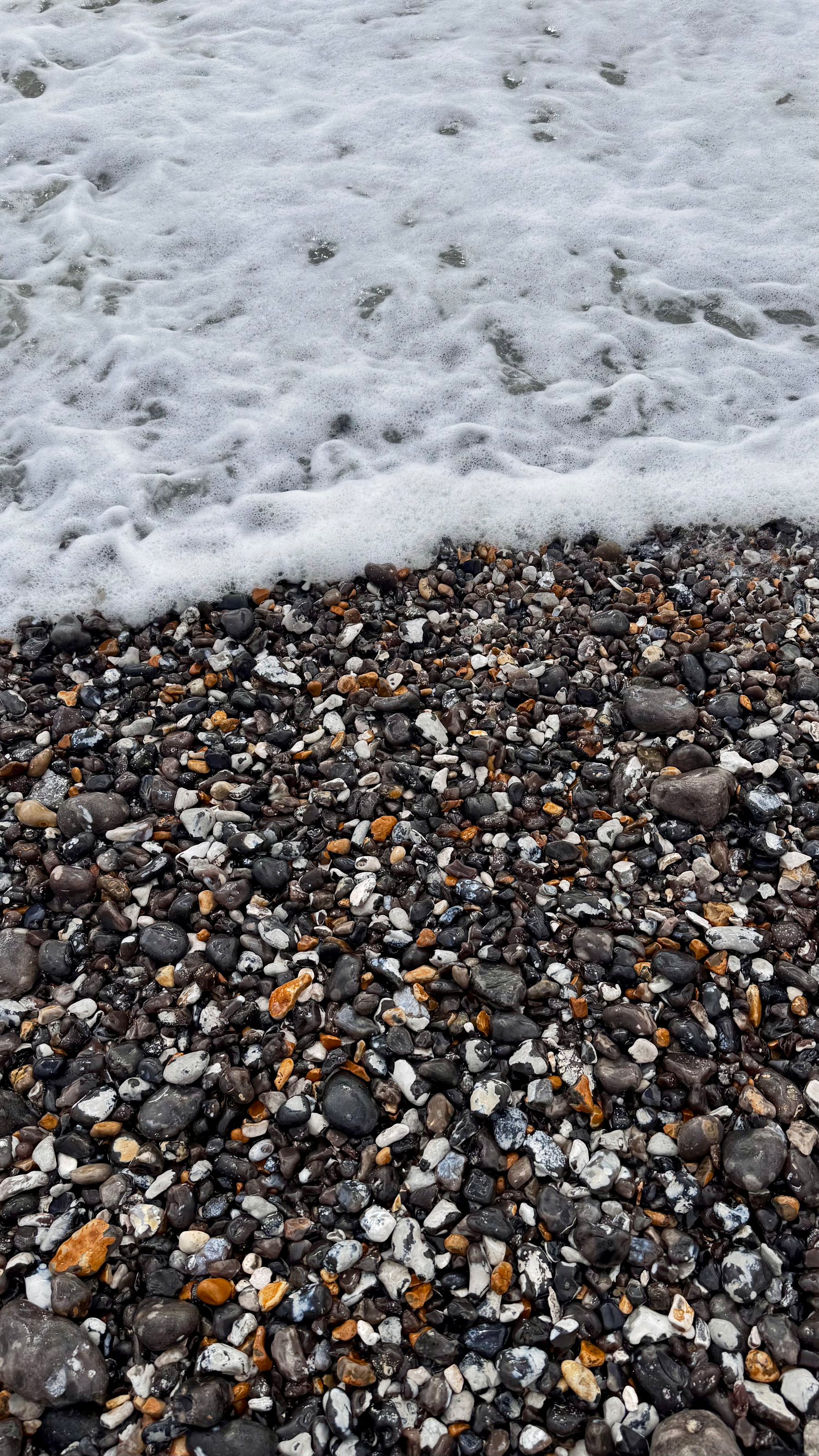 A foamy wave is gently washing over a pebble-covered shoreline with a mix of black, white, and brown stones.