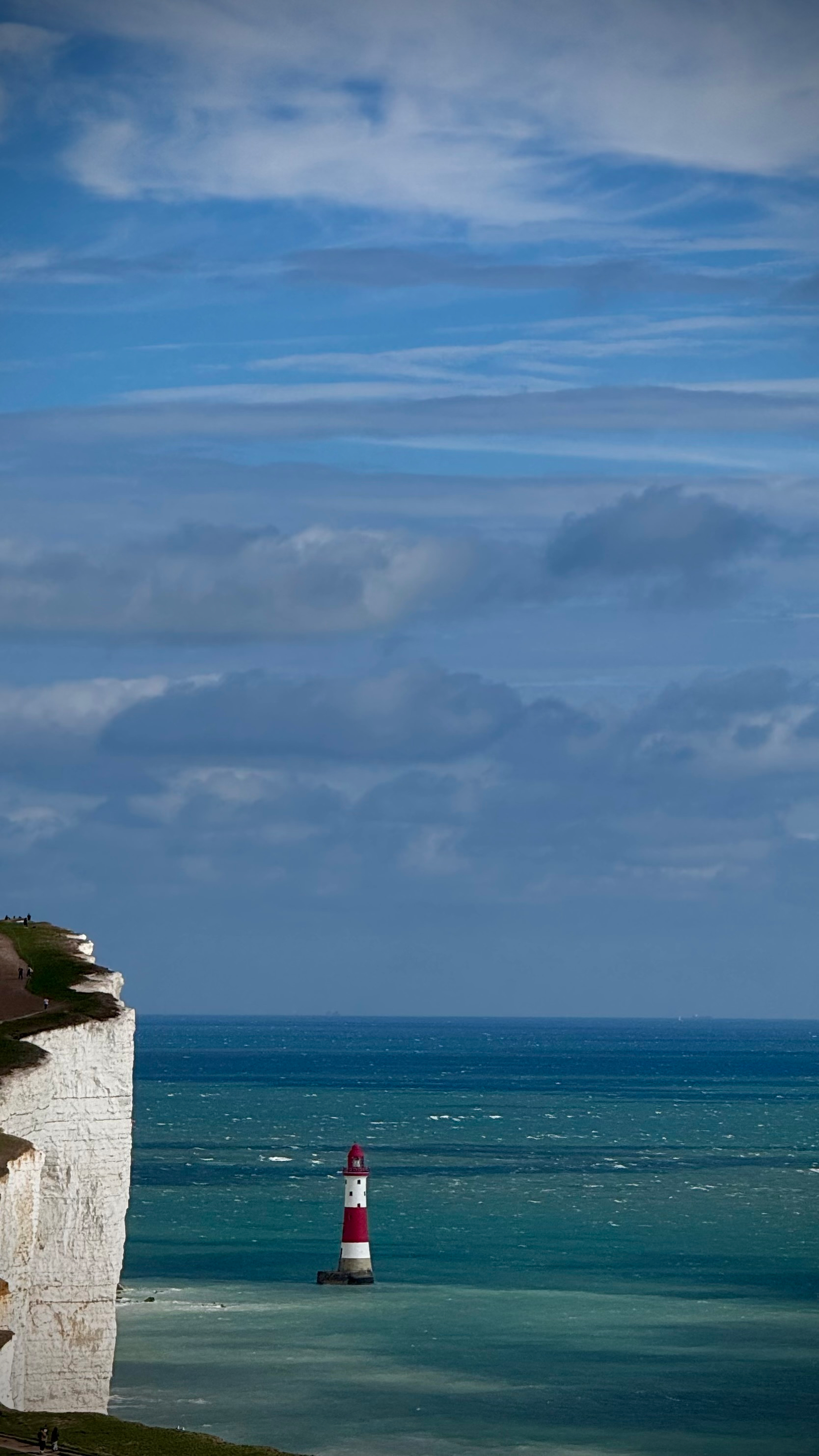 A red and white striped lighthouse stands in the sea near a white chalk cliff under a partly cloudy sky.