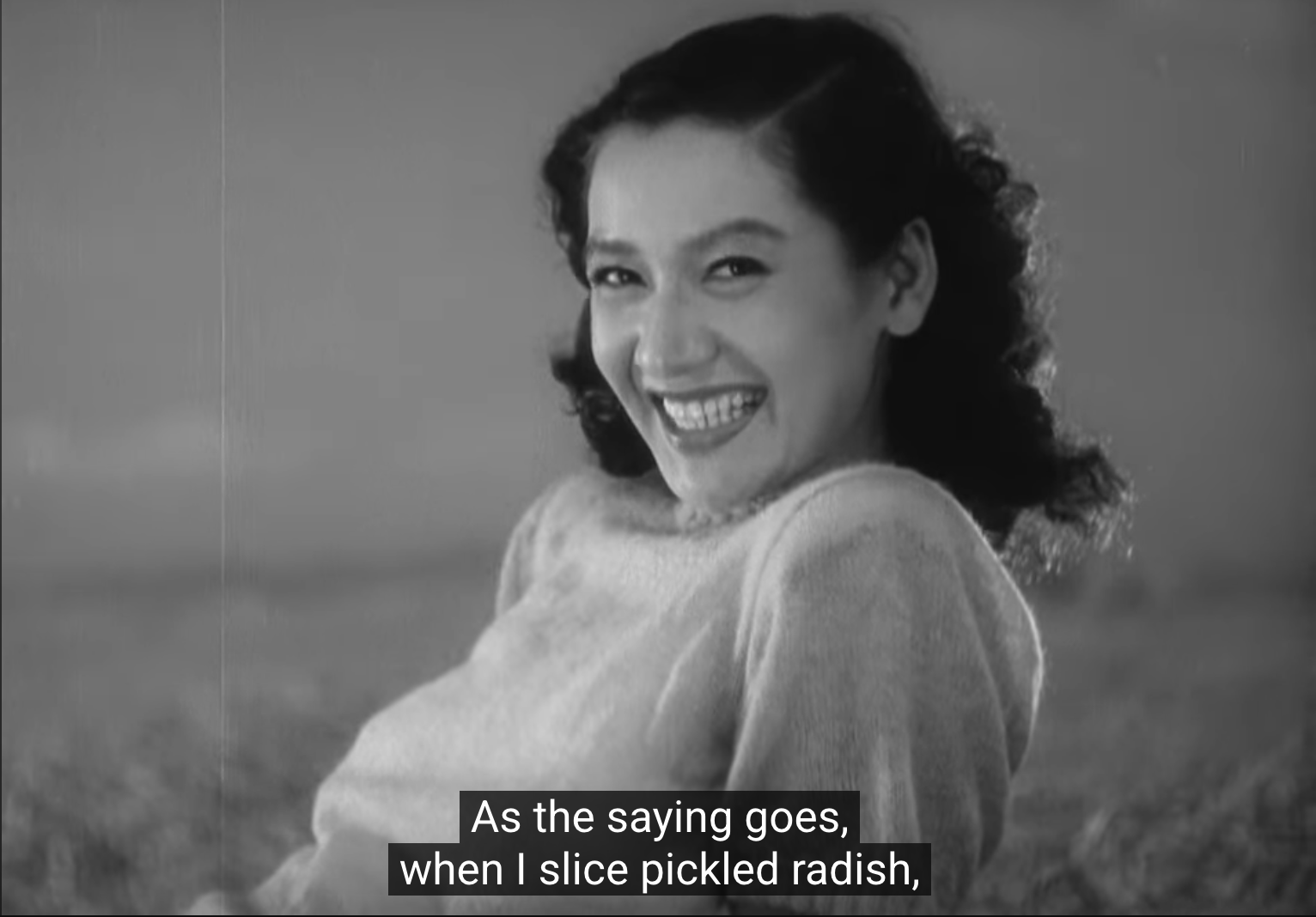 a still from ozu's "late spring" of noriko (setsuko hara) at the beach with hattori (jun usami; off-screen), where noriko is smiling and is saying "[as the saying goes,] when i sliced pickled radish ..."